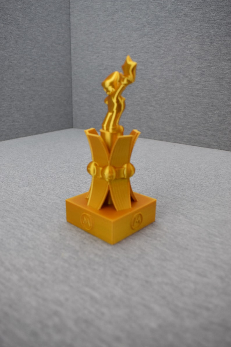 May include: A gold-colored trophy with a stylized figure on top. The base of the trophy is square with a decorative design.