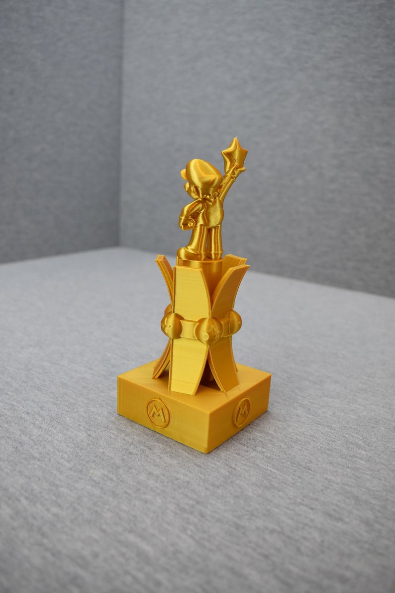 May include: A gold-colored trophy featuring a cartoon character holding a star. The trophy has a base with a circular design and the letter "M" on each side.