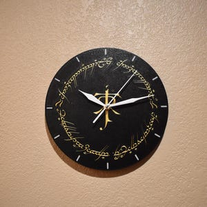 May include: A black clock with white hands and gold lettering around the edge. The lettering reads "One Ring to rule them all, One Ring to find them, One Ring to bring them all and in the darkness bind them."
