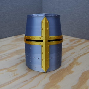 Puede incluir: Un casco plateado con un diseño de cruz dorada. El casco tiene una superficie texturizada y está decorado con pequeños remaches.