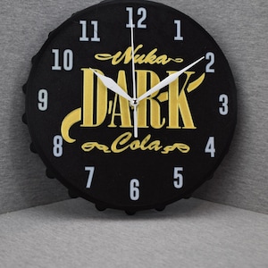 Puede incluir: Reloj de pared negro con forma de tapón de botella, con las palabras "Nuka Dark Cola" en letras doradas. El reloj tiene manecillas y números blancos. Un artículo decorativo único.