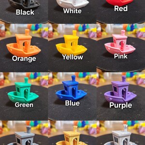 Op de afbeelding: Een set van 12 miniatuur speelgoedbootjes in verschillende kleuren, waaronder zwart, wit, rood, oranje, geel, roze, groen, blauw, paars, grijs, goud en zilver. Elk bootje heeft een kleine schoorsteen en een eenvoudig ontwerp.