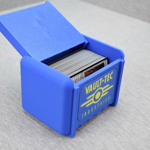以下が含まれることがあります： 黄色のVault-Tec Industriesのロゴがついた青いプラスチック製のカード収納ボックス。ボックスは開いており、カードでいっぱいです。