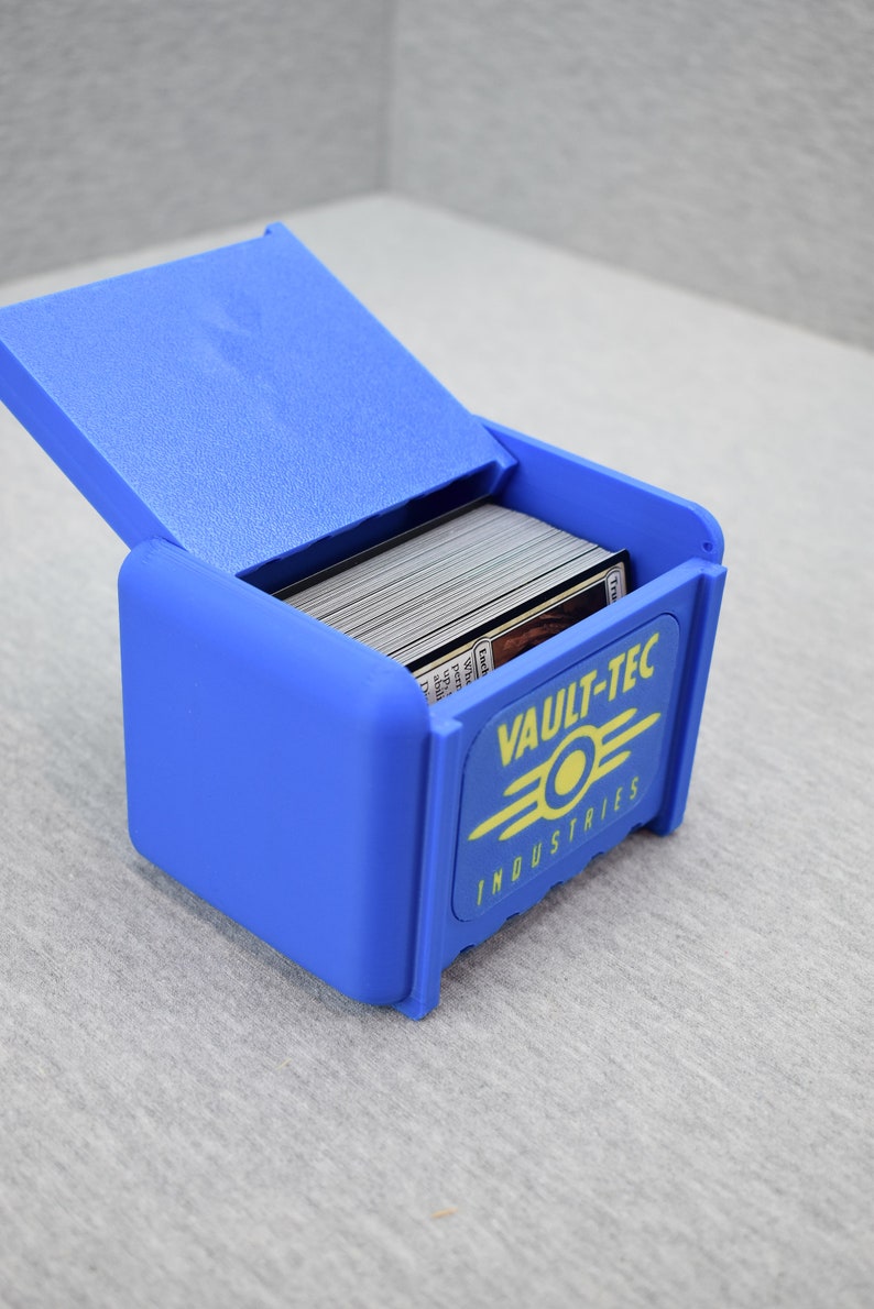 以下が含まれることがあります： 黄色のVault-Tec Industriesのロゴがついた青いプラスチック製のカード収納ボックス。ボックスは開いており、カードでいっぱいです。
