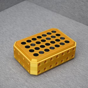Op de afbeelding: Een gouden plastic opbergdoos met een raster van ronde gaten. De gaten zijn gevuld met zwarte cirkels.