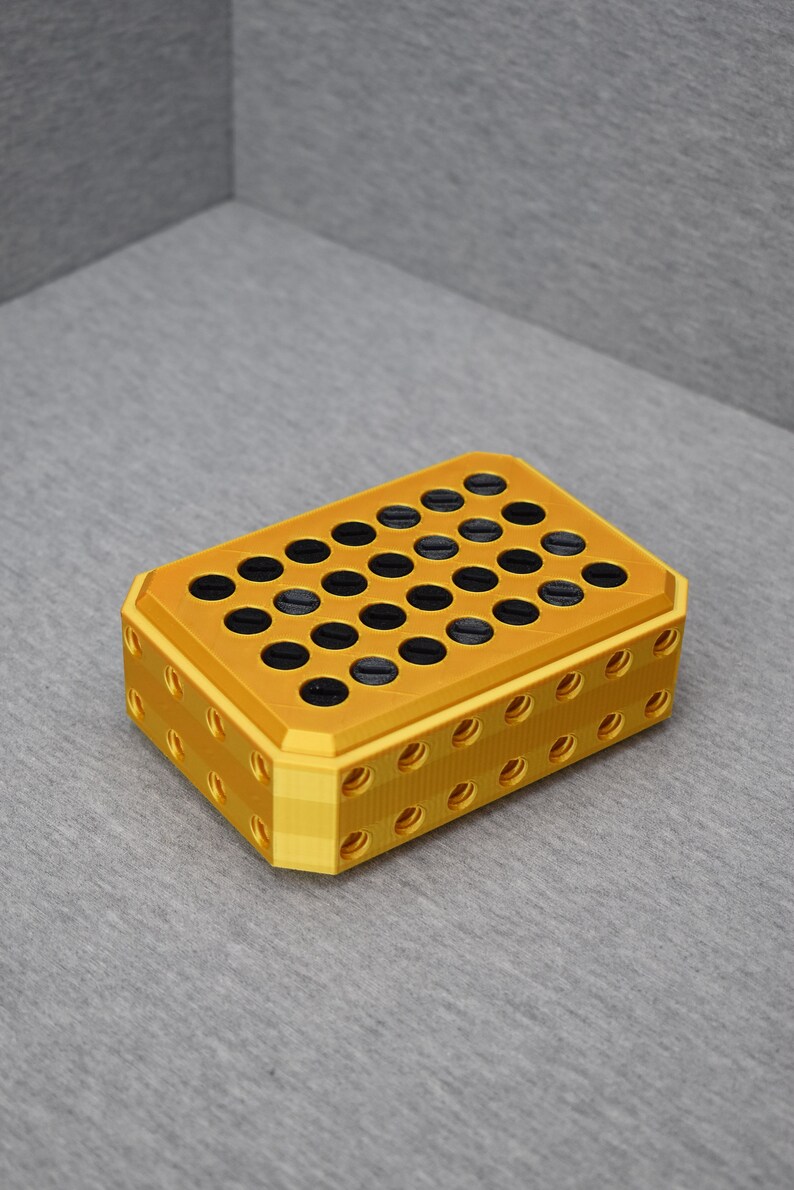 Op de afbeelding: Een gouden plastic opbergdoos met een raster van ronde gaten. De gaten zijn gevuld met zwarte cirkels.