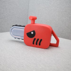 Op de afbeelding: Een rood speelgoedkettingzaag met een zwart-wit gezichtsontwerp. De kettingzaag heeft een zilveren mes en een handvat. Het speelgoed is ontworpen om eruit te zien als een stripfiguur.