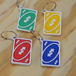 Puede incluir: Cuatro llaveros con la carta "Reverse" del juego de cartas Uno. Cada llavero tiene un color diferente: verde, amarillo, rojo y azul. Las cartas tienen un borde blanco y un llavero plateado.