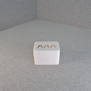May include: A white, rectangular box with the letters "AAA" cut out on the top. The box is sitting on a gray surface. The box has a simple, minimalist design.