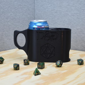 Puede incluir: Taza negra impresa en 3D con asa, diseñada para contener una lata de refresco. La taza presenta el logotipo de Dungeons & Dragons y un diseño de dado de 20 caras. Una lata azul de agua con gas está dentro de la taza y varios dados están esparcidos.
