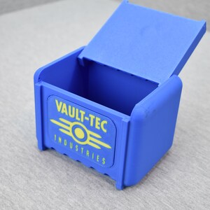 以下が含まれることがあります： 蓋付きの青いプラスチック製の箱。箱の前面には、黄色と白のVault-Tec Industriesのロゴがあります。