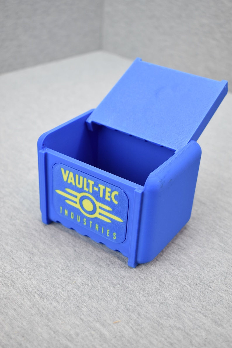 以下が含まれることがあります： 蓋付きの青いプラスチック製の箱。箱の前面には、黄色と白のVault-Tec Industriesのロゴがあります。