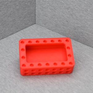 Op de afbeelding: Een rode siliconen bak met een rechthoekige vorm en een verhoogde rand. De bak heeft een reeks kleine, gelijkmatig verdeelde gaten rond de omtrek en een grotere rechthoekige opening in het midden.