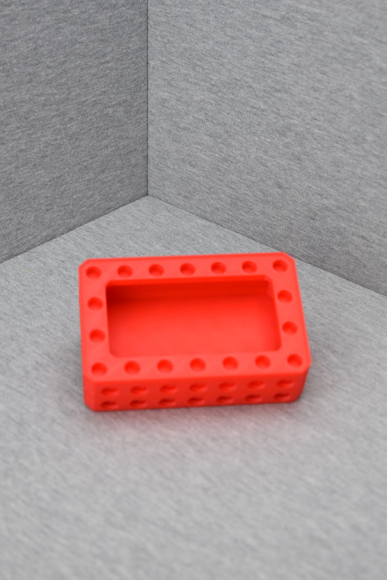 Op de afbeelding: Een rode siliconen bak met een rechthoekige vorm en een verhoogde rand. De bak heeft een reeks kleine, gelijkmatig verdeelde gaten rond de omtrek en een grotere rechthoekige opening in het midden.