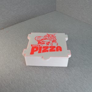 May include: A white pizza box with red text and graphics. The box features the words "NINJA PIZZA" in red, along with a cartoon graphic. The box is rectangular with a hinged lid.
