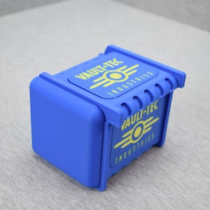 以下が含まれることがあります： 上面と側面に黄色のVault-Tec Industriesのロゴがついた青いプラスチック製の箱。箱はヒンジ付きの蓋とテクスチャ加工された表面を持っています。