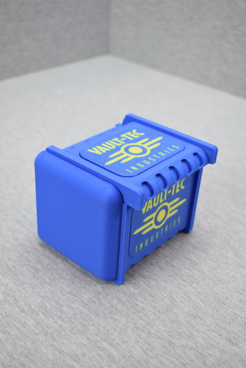 以下が含まれることがあります： 上面と側面に黄色のVault-Tec Industriesのロゴがついた青いプラスチック製の箱。箱はヒンジ付きの蓋とテクスチャ加工された表面を持っています。