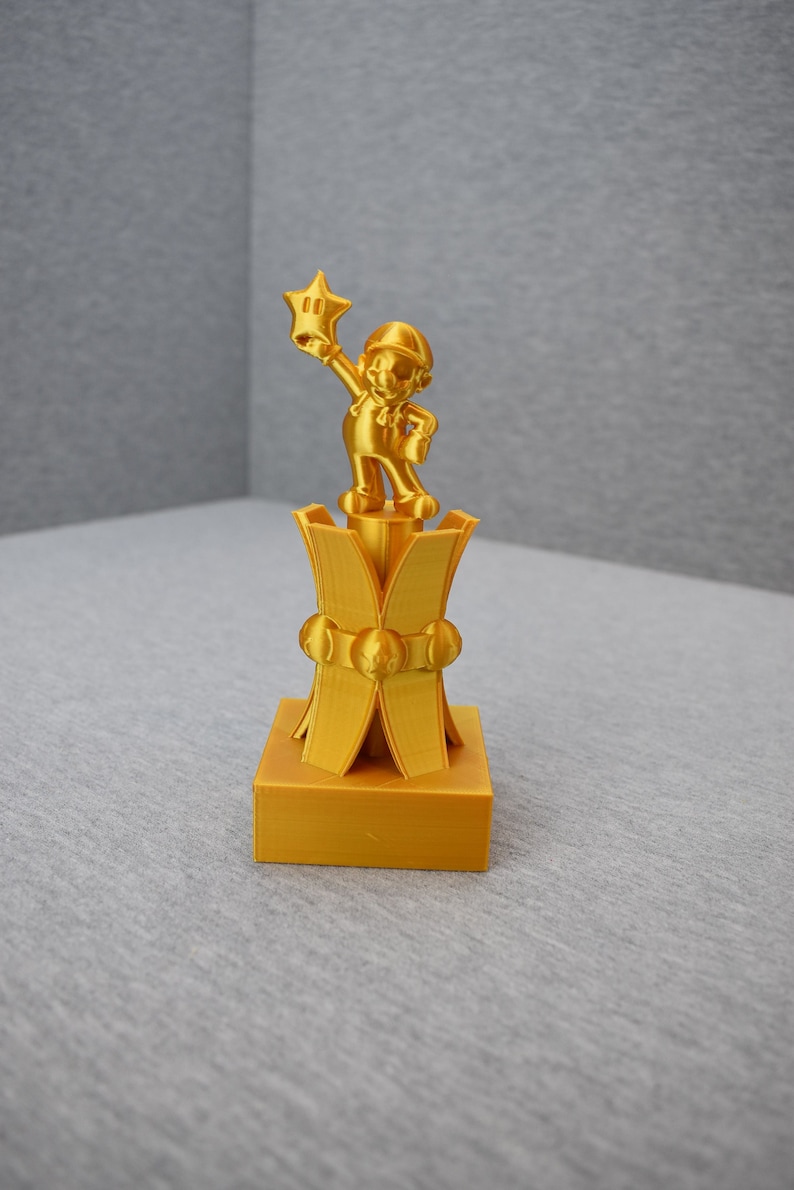 May include: A gold-colored 3D printed trophy featuring a character holding a star. The trophy has a square base and a decorative top with a star-shaped design.