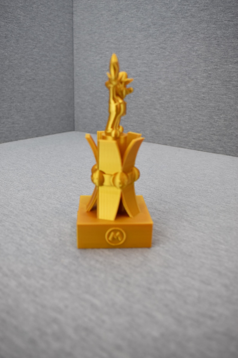 May include: A gold-colored trophy with a stylized figure on top and a circular emblem with the letter "M" on the base.