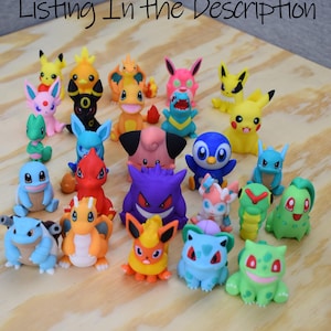 May include: A collection of colorful Pokémon figurines, including Pikachu, Charmander, and Bulbasaur, are arranged on a light wood surface. The figurines are various sizes and colors, with the text "More Pokemon On My other Listing In the Description" at the top.