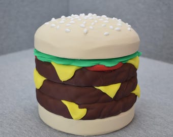 Może przedstawiać: Duży, okrągły, trzywarstwowy hamburger z plasteliny. Hamburger jest przykryty bułką z sezamem, sałatą, pomidorem i serem.