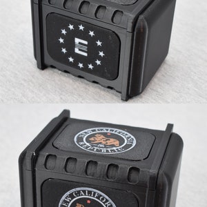 以下が含まれることがあります： 上部に異なるデザインが施された黒いプラスチック製の箱が2つあります。最初の箱は、星と「E」の文字が描かれた白黒のデザインです。2番目の箱は、クマと「NEW CALIFORNIA REPUBLIC」の文字が描かれた茶色と白のデザインです。