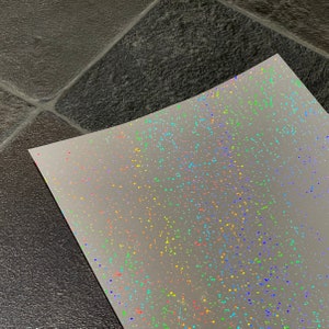 May include: A sheet of iridescent holographic paper with a scattered pattern of rainbow glitter.