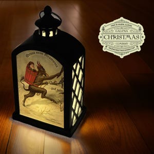 May include: A black lantern with a white image of Krampus, a mythical creature, carrying a child on his back. The lantern has a white inscription that reads "Gruss vom Krampus" and "gut sein". The lantern is made by Galena Christmas Company.