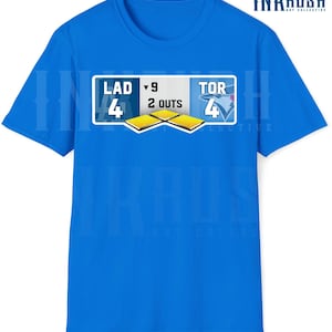 Peut inclure: Un t-shirt bleu avec un motif de tableau de bord de baseball. Le tableau de bord affiche les scores pour LAD et TOR, avec le texte "9 2 OUTS". Le t-shirt porte également le logo "INKRUSH ART COLLECTIVE".