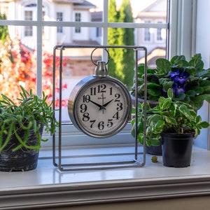 Pode incluir: Um relógio prateado com mostrador branco e algarismos pretos está numa janela, emoldurado por um suporte metálico retangular. Plantas em vasos com folhas verdes e flores roxas estão ao lado, com uma janela e árvores ao fundo.