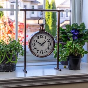 Pode incluir: Um relógio decorativo de cor bronze com mostrador branco e algarismos pretos está pendurado em um suporte de metal. Plantas em vasos com folhas verdes e flores roxas estão em uma janela branca. O fundo mostra uma janela com vista para árvores.