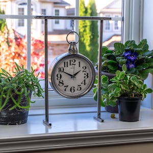 Pode incluir: Um relógio prateado em estilo vintage, com mostrador branco e algarismos pretos, pende de um suporte de metal. O relógio é exibido numa janela, juntamente com plantas em vasos com folhagem verde e flores roxas. O fundo mostra árvores e casas.