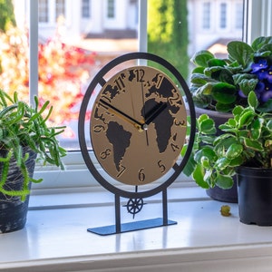 Puede incluir: Un reloj de escritorio decorativo con un diseño de mapa del mundo. La esfera del reloj es dorada con continentes negros y manecillas negras. El reloj está enmarcado en un marco y base de metal negro. El reloj se exhibe en un alféizar blanco.