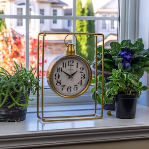 Pode incluir: Um relógio de metal dourado com mostrador branco e números pretos, emoldurado em uma moldura retangular. O relógio está em uma janela, ladeado por plantas em vasos com folhagem verde e flores roxas. O relógio diz "Galaxy".