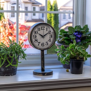 May include: A vintage-style clock with a black and white face stands on a windowsill, flanked by potted plants. The clock's hands show the time. The window reveals a view of trees and a house in the background.