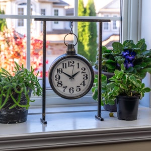 Pode incluir: Um relógio decorativo com moldura preta e mostrador branco está pendurado em um suporte de metal. O relógio é cercado por plantas em vasos com folhagem verde e flores roxas, colocadas em uma janela. O fundo mostra uma vista de uma casa e árvores.