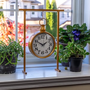Pode incluir: Um relógio dourado em estilo vintage com mostrador branco e algarismos pretos pende de um suporte a condizer. O relógio é exibido numa janela, ladeado por plantas em vasos com folhagem verde e flores roxas. O fundo mostra uma casa e árvores.