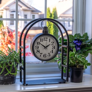 Pode incluir: Um relógio de metal preto com mostrador branco e algarismos pretos está numa janela. O relógio é suportado por um suporte decorativo. Plantas em vasos, incluindo uma suculenta e uma violeta africana, ladeiam o relógio. O fundo mostra casas e árvores.