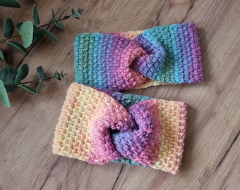May include: Two handmade crochet headbands in a rainbow color scheme. The headbands feature a twisted knot design and are made with a soft, textured yarn. The colors transition from yellow to pink, purple, and blue.