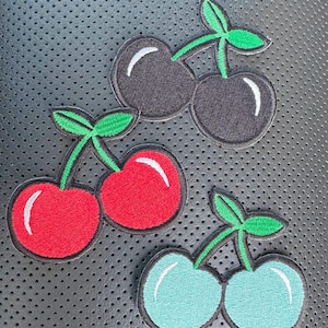 May include: Embroidered cherry patches in red, black, and teal with green stems and leaves. Each patch features a pair of cherries with white highlights. The patches are set against a dark, textured background.