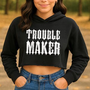 May include: Black cropped hoodie with the text "TROUBLE MAKER" in white, gothic-style lettering. The hoodie has a drawstring hood and long sleeves. The model is wearing blue jeans.