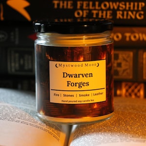 May include: A glass jar candle with a black lid, labeled "Dwarven Forges" by Mystwood Moon. The label lists "Fire, Stones, Smoke, Leather" as scents. The candle is 9 oz and sits on an open book, with "The Fellowship of the Ring" visible in the background.