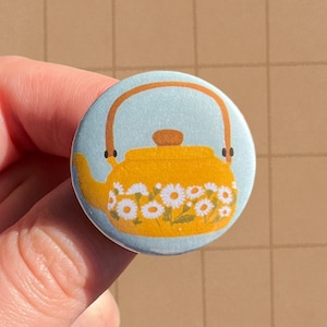 May include: A yellow teapot with a white daisy pattern on a blue background. The teapot has a brown handle and a brown lid.