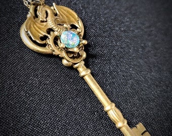 Puede incluir: Un collar con colgante en forma de llave de color latón. La cabeza de la llave presenta un diseño de dragón y una piedra preciosa ovalada iridiscente. La llave cuelga de una fina cadena. El fondo es una tela oscura y texturizada.