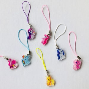 May include: Eight colorful My Little Pony character phone charms with different colored strings. The charms are made of clear plastic with a printed image of a pony inside.