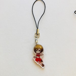 Peut inclure: Un charm en acrylique transparent repr&eacute;sentant un personnage de dessin anim&eacute; v&ecirc;tu d'une tenue rouge et aux cheveux bruns. Le charm est attach&eacute; &agrave; un cordon noir avec un fermoir en m&eacute;tal argent&eacute;. Le fond est une surface blanche unie.