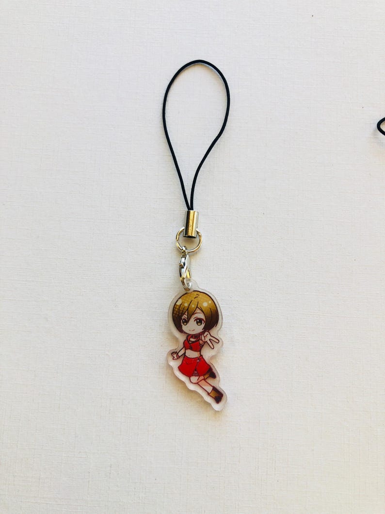 Peut inclure: Un charm en acrylique transparent repr&eacute;sentant un personnage de dessin anim&eacute; v&ecirc;tu d'une tenue rouge et aux cheveux bruns. Le charm est attach&eacute; &agrave; un cordon noir avec un fermoir en m&eacute;tal argent&eacute;. Le fond est une surface blanche unie.