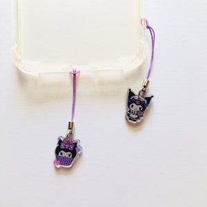 Op de afbeelding: Twee doorzichtige telefoonbedels met paarse lanyards. E&eacute;n bedel heeft een stripfiguur in een cupcake, de andere een figuur met een schedelontwerp. De bedels zijn bevestigd aan een doorzichtige telefoonhoes.