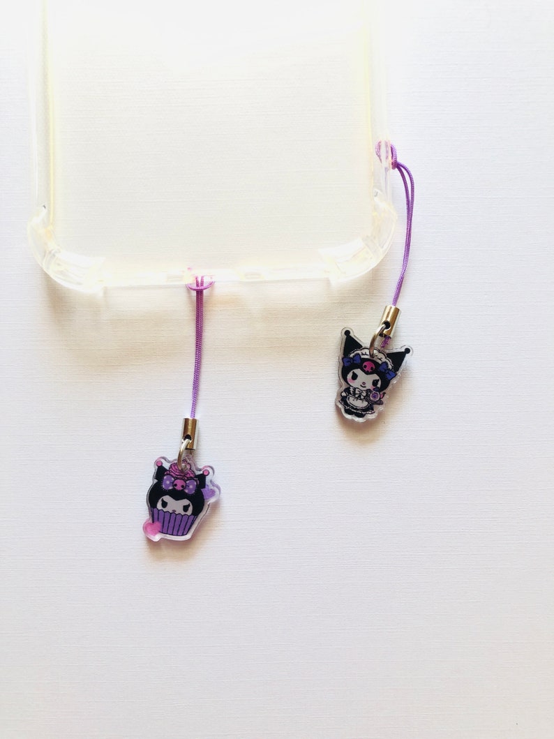 Op de afbeelding: Twee doorzichtige telefoonbedels met paarse lanyards. E&eacute;n bedel heeft een stripfiguur in een cupcake, de andere een figuur met een schedelontwerp. De bedels zijn bevestigd aan een doorzichtige telefoonhoes.
