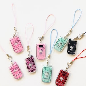 May include: A collection of eight acrylic phone charms featuring Hello Kitty designs on miniature flip phone shapes. Each charm includes a colorful lanyard strap and metal clasp, showcasing various patterns like leopard print and solid colors.